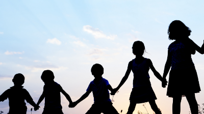 A group young children stand in a line while holding hands and appearing to be playing. They have their backs to us and are silhouetted against the sky.