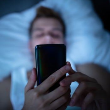 A blurred image shows a young man in a white vest lying down on a bed while holding a mobile phone up above him.