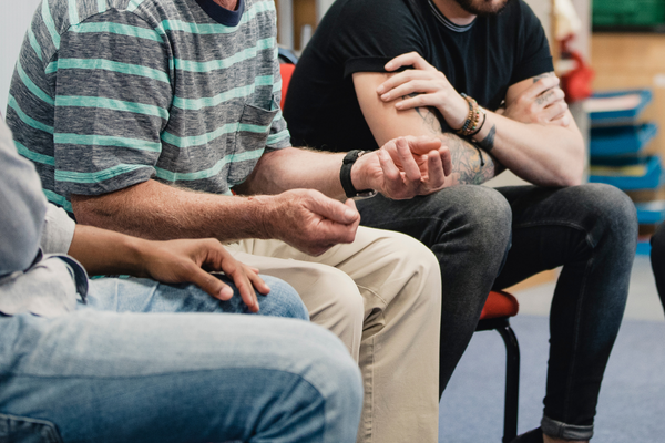 A group of men of different backgrounds are sitting in a circle. Only their bodies are visible from the shoulders down.