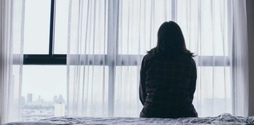 This black and white image shows the back of a woman, sitting on a bed looking out the window.