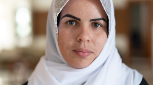 A woman wearing a white headscarf looks straight into the camera. Only her head and shoulders are visible.
