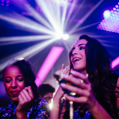 A young woman dances in a nightclub next to her friend who is holding a cocktail.