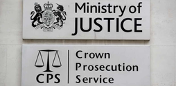 A white sign for the Ministry of Justice with a black coat of arms illustration next to it. Underneath this sign is a white sign with a black illustration of the scale of justice next to black writing that reads Crown Prosecution Service.