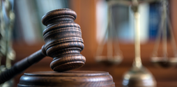 A zoomed in photo of a wooden gavel on a desk. In the background out of focus is a gold scale of  justice statue.