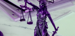 A blue tinted image of a small metal statue of Lady Justice on top of a desk. In the background of the shot is a pile of notebooks.