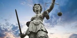 A stone statue of Lady Justice, without a blindfold on. Behind her the sun is beginning to set.