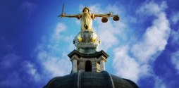 A gold statue of Lady Justice on top of a building. The photo is taken at an angle so we are looking up at Lady Justice. The sky behind the statue is blue with a few clouds.