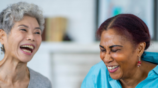 Two older women sit at a table with tea cups on it and are laughing together.