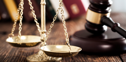 On a wooden desk rests a silver scale of justice which is in shot. There is a blurred gavel behind the scales.