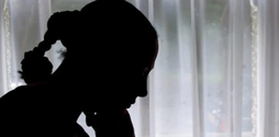 A woman rests her chin on her hand and looks down sadly. She is sat by a window, so we can not see her face just her silhouette.